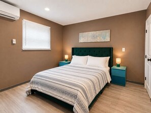 A bedroom with a bed, two bedside tables with lamps, a window with blinds, and a wall-mounted air conditioner.