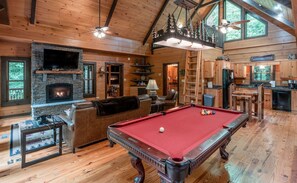 Interior acogedor de una cabaña con mesa de billar, chimenea, piso de madera y zona de cocina.