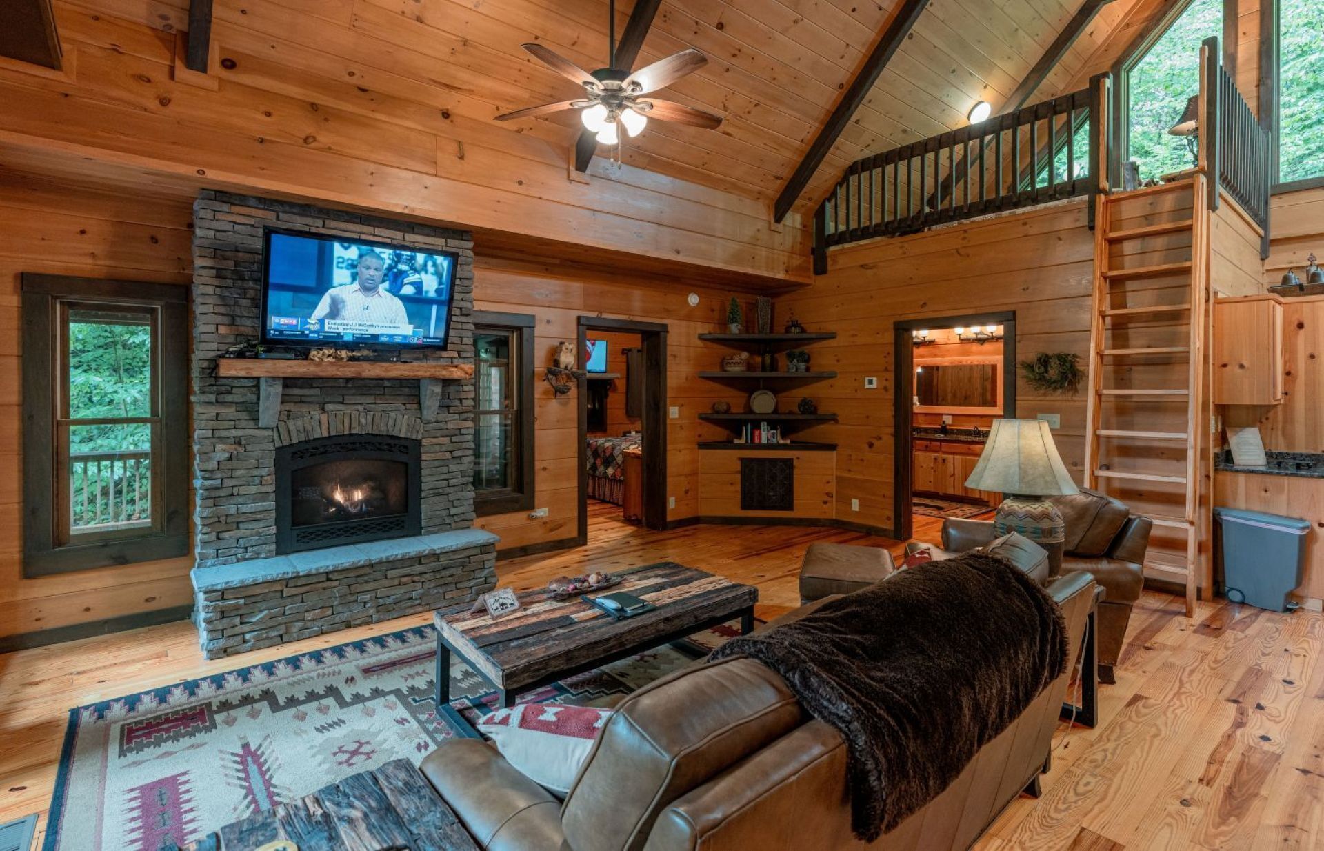 A cozy cabin interior with a stone fireplace, wooden walls, a television, and a dining area.