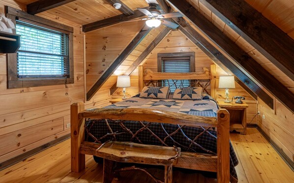 Un intérieur en bois avec un plafond en pente, un lit recouvert d’une couvre-lit à motifs géométriques, deux lampes de chevet et une fenêtre avec des stores.