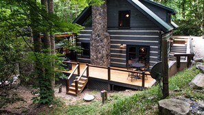 A cabin with a stone chimney, wooden deck, and outdoor seating area surrounded by trees. - Water's Edge-Pet Friendly 3 Bedroom, Luxury Log Cabin Rental, Perfect for Families, Outdoor Entertaining, Hot Tub, Fireplace, 30 Minutes to Great Smoky Mountains-Nantahala NC (Topton)