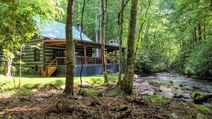 A log cabin with a metal roof, surrounded by a forest and a stream. - Water's Edge-Pet Friendly 3 Bedroom, Luxury Log Cabin Rental, Perfect for Families, Outdoor Entertaining, Hot Tub, Fireplace, 30 Minutes to Great Smoky Mountains-Nantahala NC (Topton)