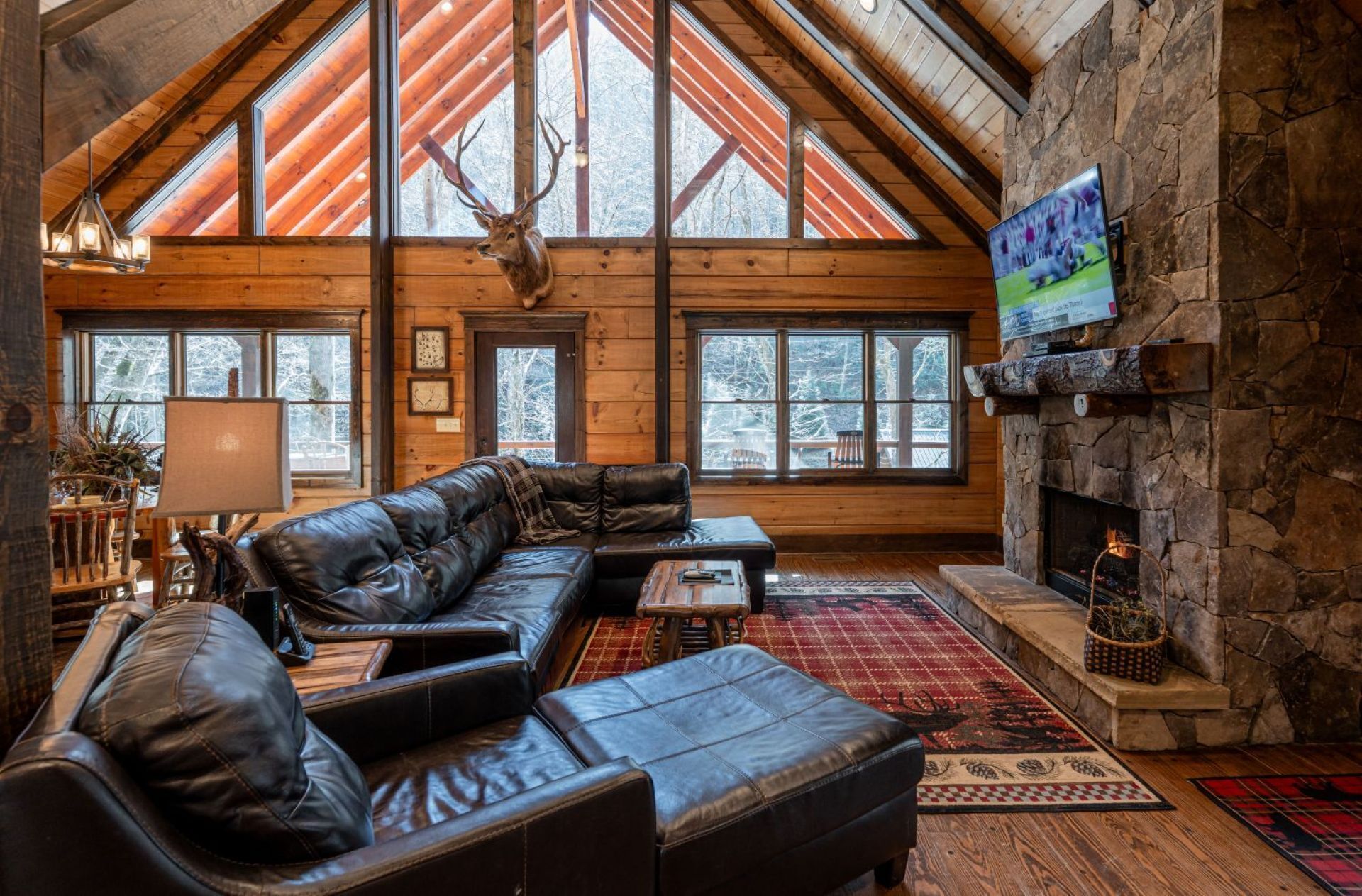 A log cabin interior with a stone fireplace, leather sofa, and large windows.