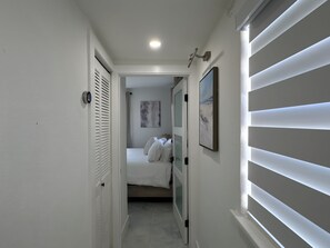 A narrow hallway leading to a bedroom with a bed, a door with a frosted glass panel, and a wall-mounted picture.