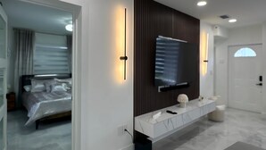 Un dormitorio moderno con cama, televisor montado en un panel oscuro, una mesita consola de mármol y un baño con puerta blanca.