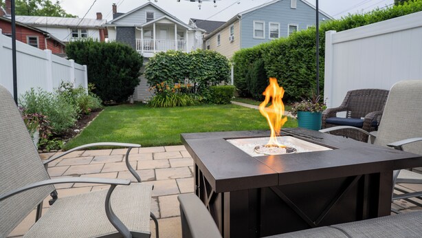 An outdoor patio with a square table and four chairs, a fire pit with flames, a well-maintained lawn, and a white fence. - Stylish Apt Walk to Shops, Dining and Bucknell (Lewisburg)