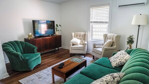 A living room with a wooden coffee table, two green sofas, two beige armchairs, a TV on a wooden cabinet, and a window with blinds. - Stylish Apt Walk to Shops, Dining and Bucknell (Lewisburg)