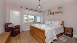 A bedroom with a wooden bed, a chair, a bedside table, a window with curtains, and a hanging light fixture. - Coastal Charm In Clacton On Sea / With Balcony & Sea View / Sleeps 4 (Essex)