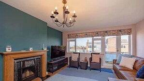 A living room with a fireplace, television, sofa, and a large window with a view of buildings. - Coastal Charm In Clacton On Sea / With Balcony & Sea View / Sleeps 4 (Essex)