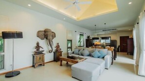 A spacious living room with a ceiling fan, a sofa set, a coffee table, and decorative items including a Buddha statue and an elephant head. - Ivory Pool Villa in Nai Harn (Phuket)