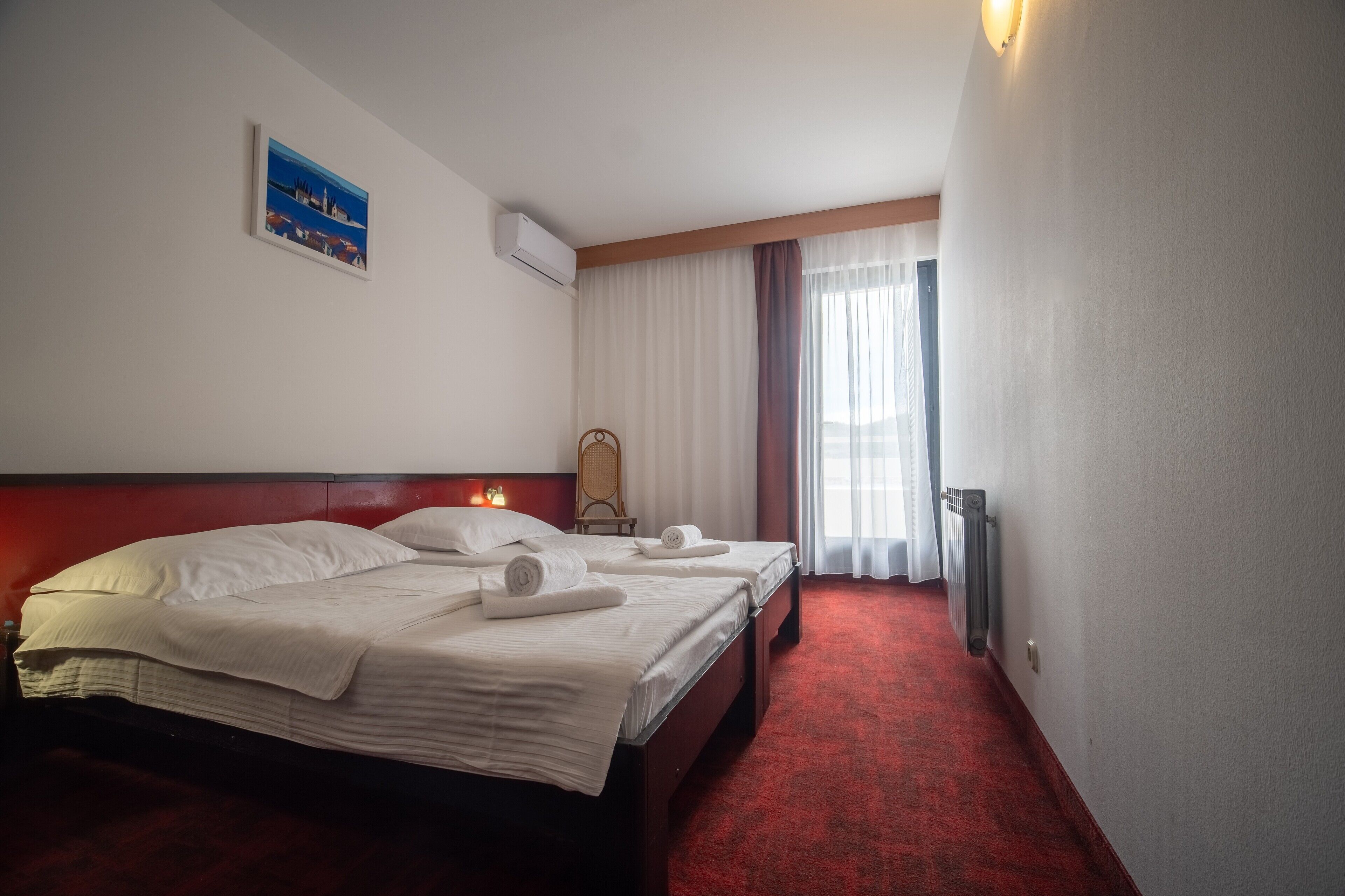 A hotel room with a large bed, a small table, a chair, a radiator, and a window with curtains.