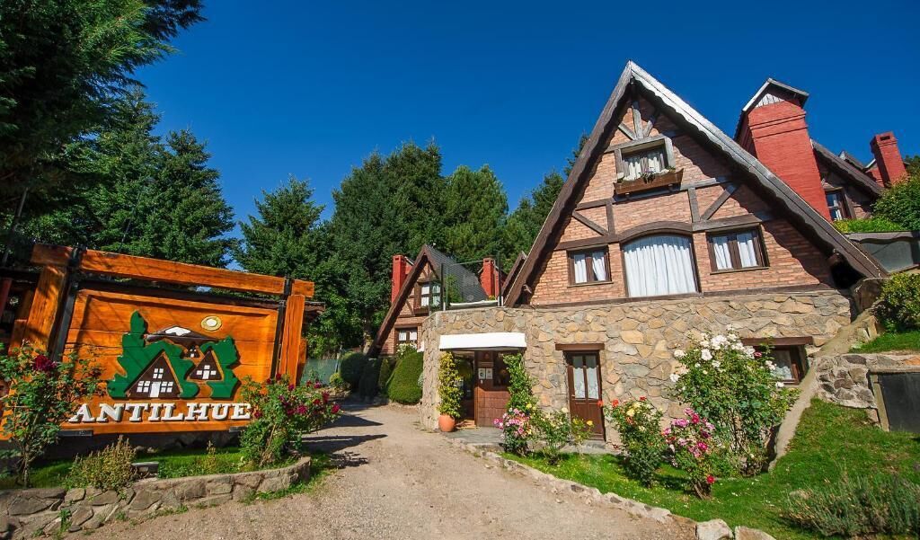 A traditional stone house with a steeply pitched roof, a wooden sign with 'ANTILHUE' written on it, and a well-maintained garden with flowers and shrubs.