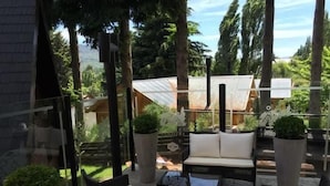 Un área de estar al aire libre con mobiliario de mimbre y plantas en macetas, con vista a un jardín y un edificio con techo metálico.