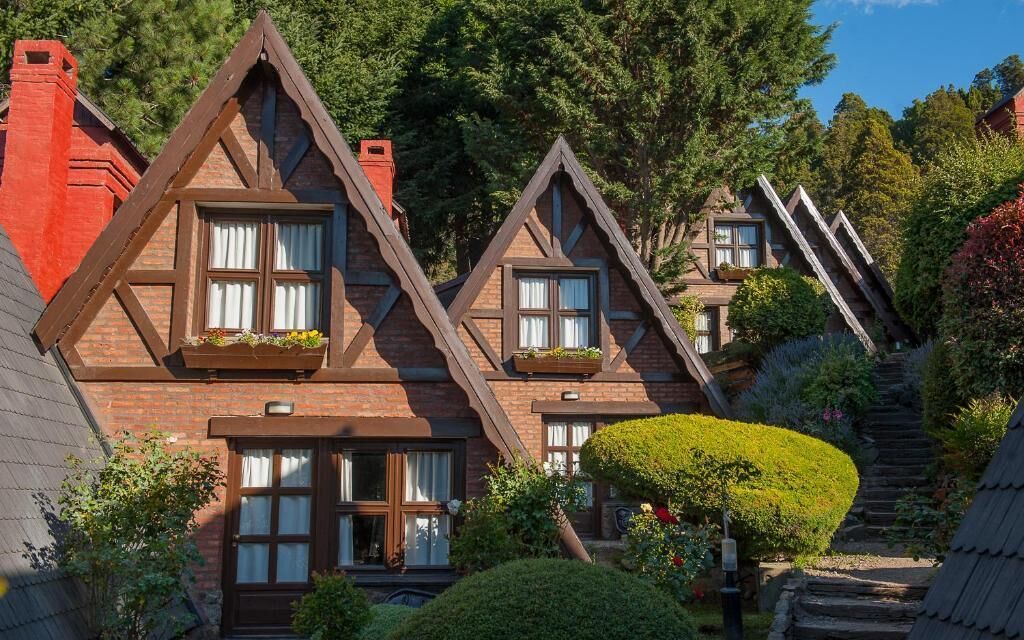 Een chalet in traditionele stijl, met steil schuin aflopende daken, dakkapellen en een verzorgde tuin met een haag en een boom.