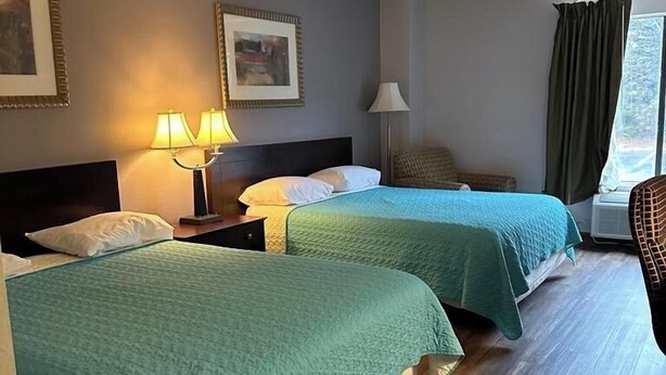 A hotel room with two beds, a nightstand with lamps, a chair, and a window with curtains. - Lawrenceville Inn (Lawrenceville)