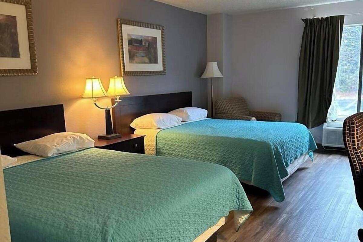 A hotel room with two beds, a nightstand with lamps, a chair, and a window with curtains.