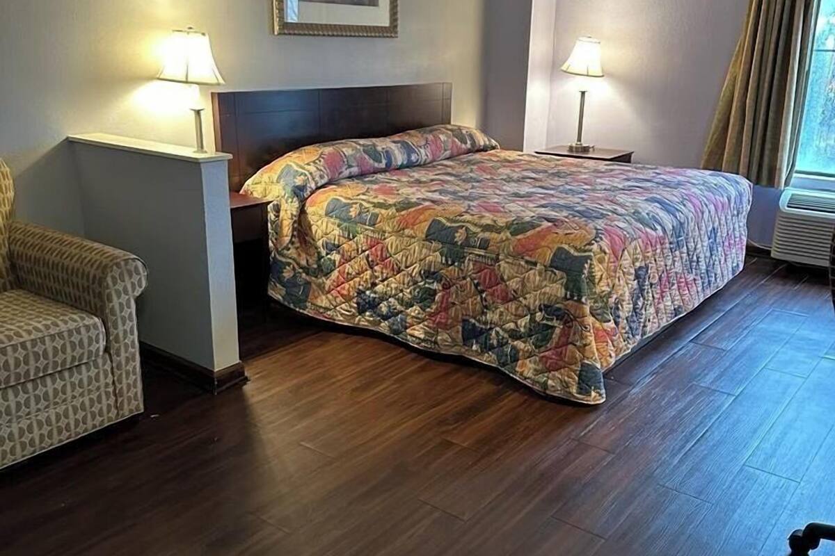 A hotel room with a bed, two bedside lamps, a chair, a framed picture on the wall, and a window with curtains.