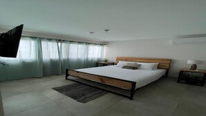 Un dormitorio con cama, mesitas de noche, televisor montado en la pared y ventana con cortinas.