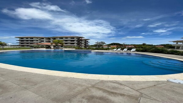 A swimming pool with a tiled deck, surrounded by a building with multiple balconies and a clear sky.