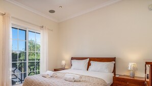 Een slaapkamer met een groot raam, een bed met witte lakens, twee bedlampen en een houten nachtkastje.