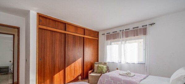 Un dormitorio con un armario de madera, una cama con colcha estampada y una ventana con cortinas translúcidas.
