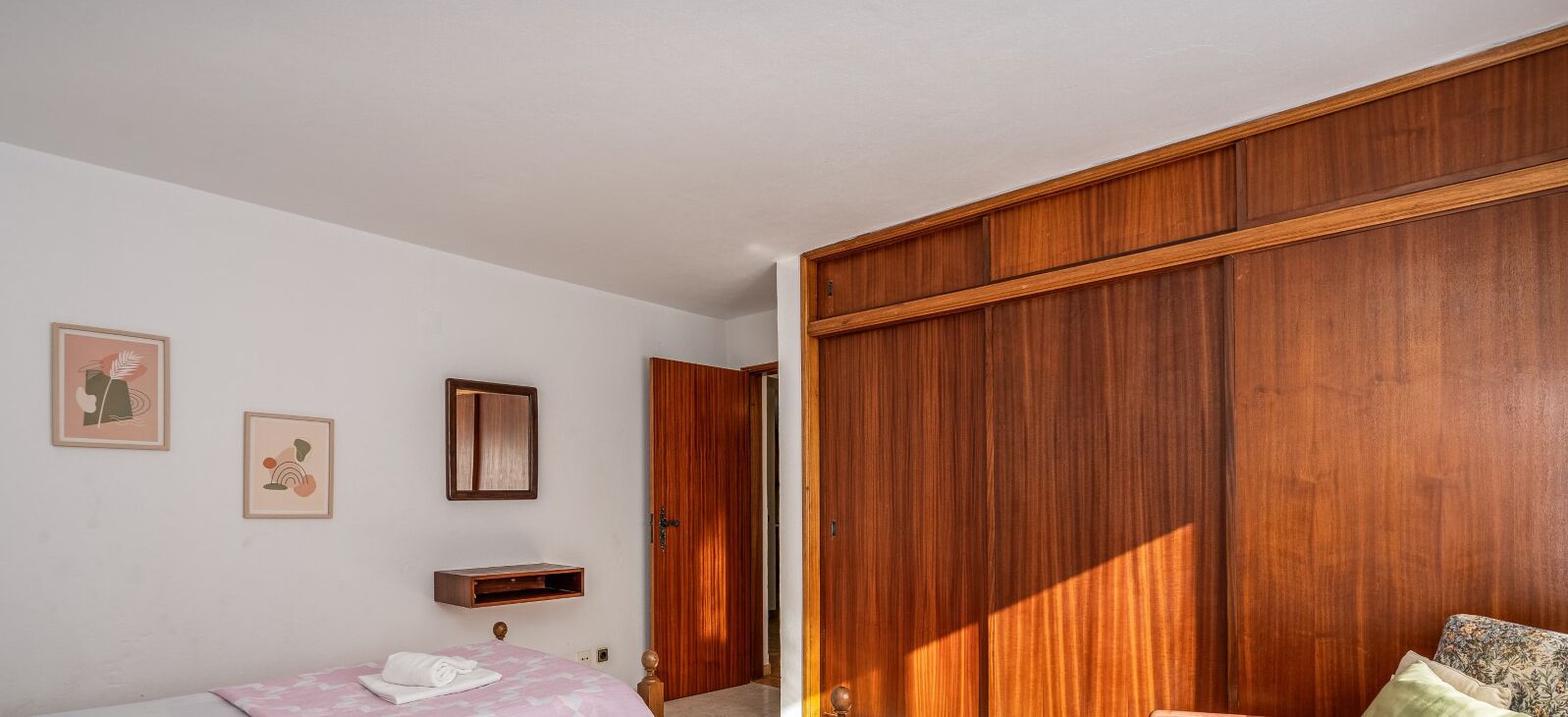 A bedroom with a wooden wardrobe, a bed with a pink bedspread, a small bedside table, and two framed pictures on the wall.