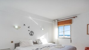 A bedroom with a bed, a window with a view of the beach, a wall-mounted light fixture, and a framed picture on the wall.