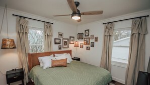 Un dormitorio con cabecera de madera, colcha estampada verde, cortinas color beige, ventilador de techo y una pared decorada con cuadros.