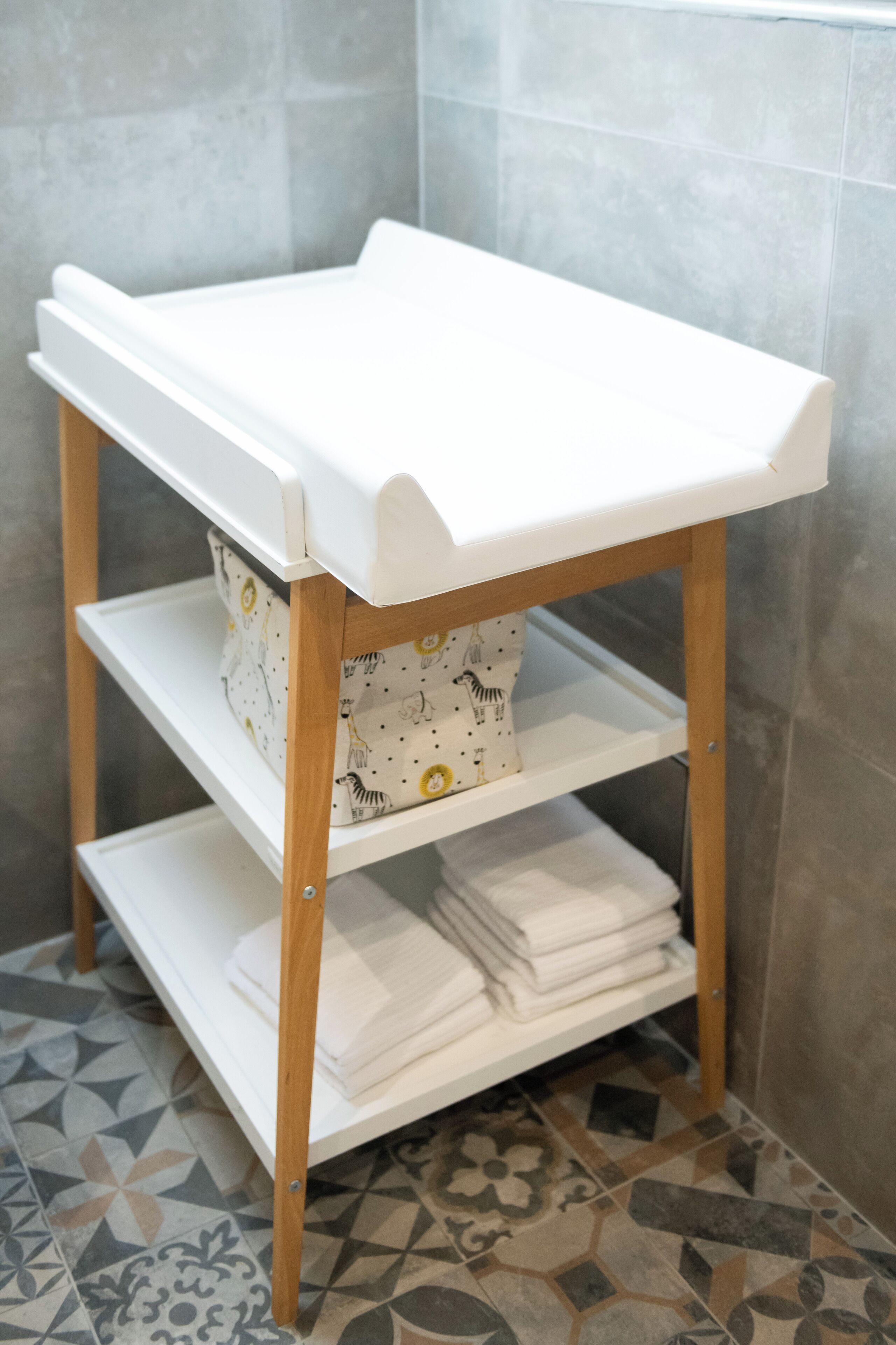 Un cambiador blanco para bebés con un estante debajo, frente a una pared de azulejos.