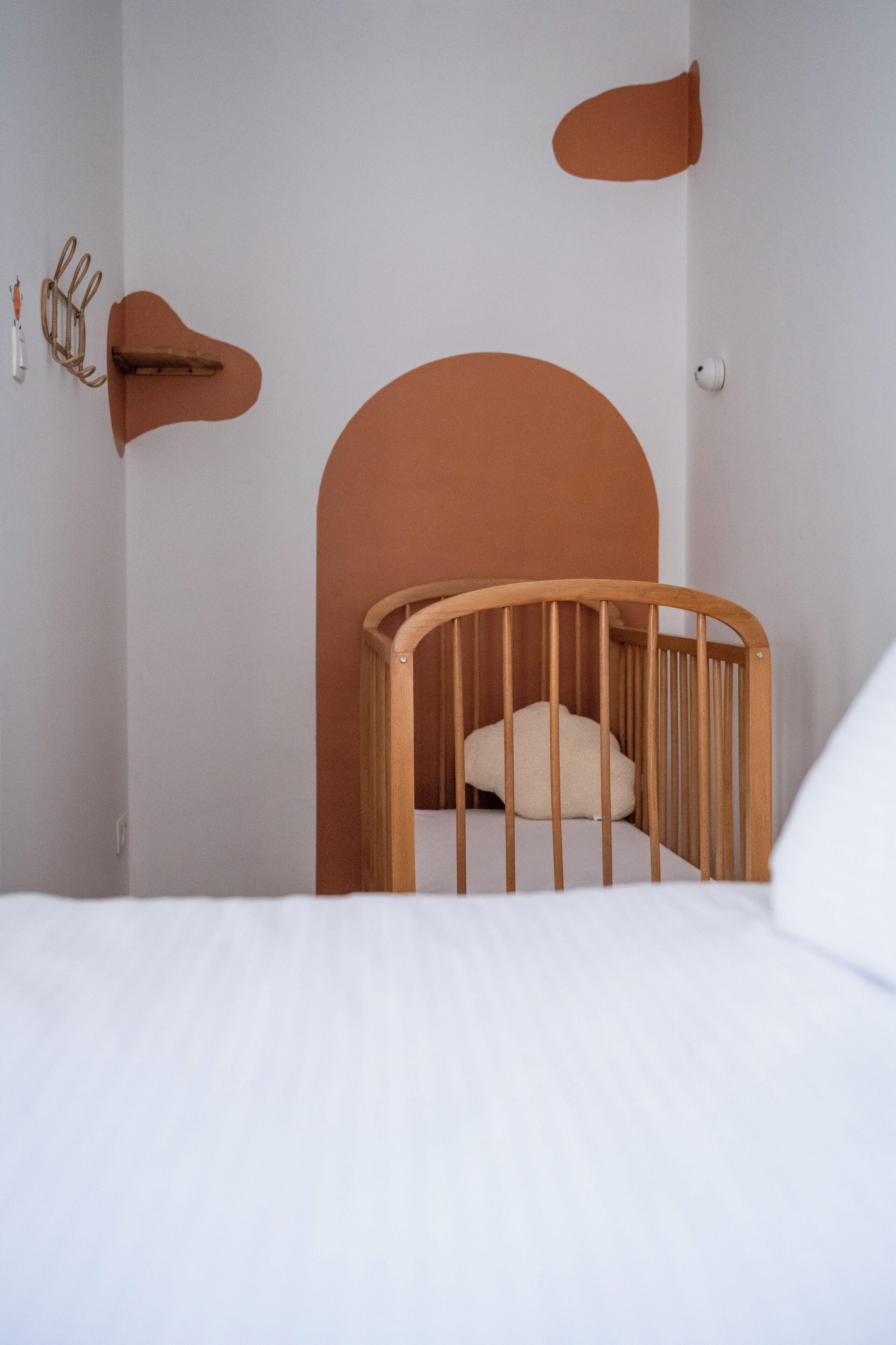 Una cuna infantil minimalista con estructura de madera curvada y un solo cojín, frente a una pared blanca lisa con un elemento decorativo naranja grande y curvo.