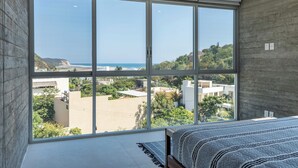 A bedroom with a large window offering a view of the ocean and surrounding buildings.