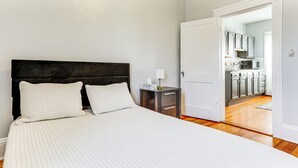 A bedroom with a large bed, a nightstand, and a view into a kitchen through an open door. - 5 Mi to Brown U: Pet-Friendly Apt, Walk to Dining! (North Providence)