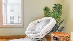 Ein Zimmer mit einem weißen Rattan-Sessel, einem runden Holzhocker und einer großen Grünpflanze.