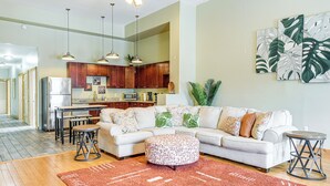 Ein modernes Wohnzimmer mit Ecksofa, Hocker und Couchtisch. Eine Küchenzeile mit Holzschränken und einer Mikrowelle ist ebenfalls vorhanden.