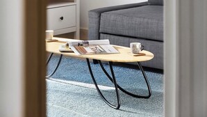 Una habitación moderna con un sofá gris, una mesa de centro de madera con revistas, una mesita de noche blanca con una lámpara y una alfombra azul.