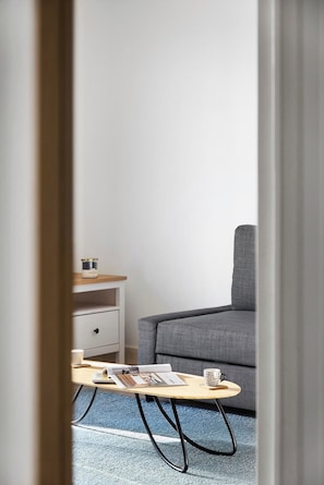 A modern room with a grey sofa, a wooden coffee table with magazines, a white bedside table with a lamp, and a blue carpet.