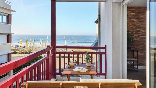 Un balcón con una mesa de madera, barandilla roja y vista a la playa y el océano.