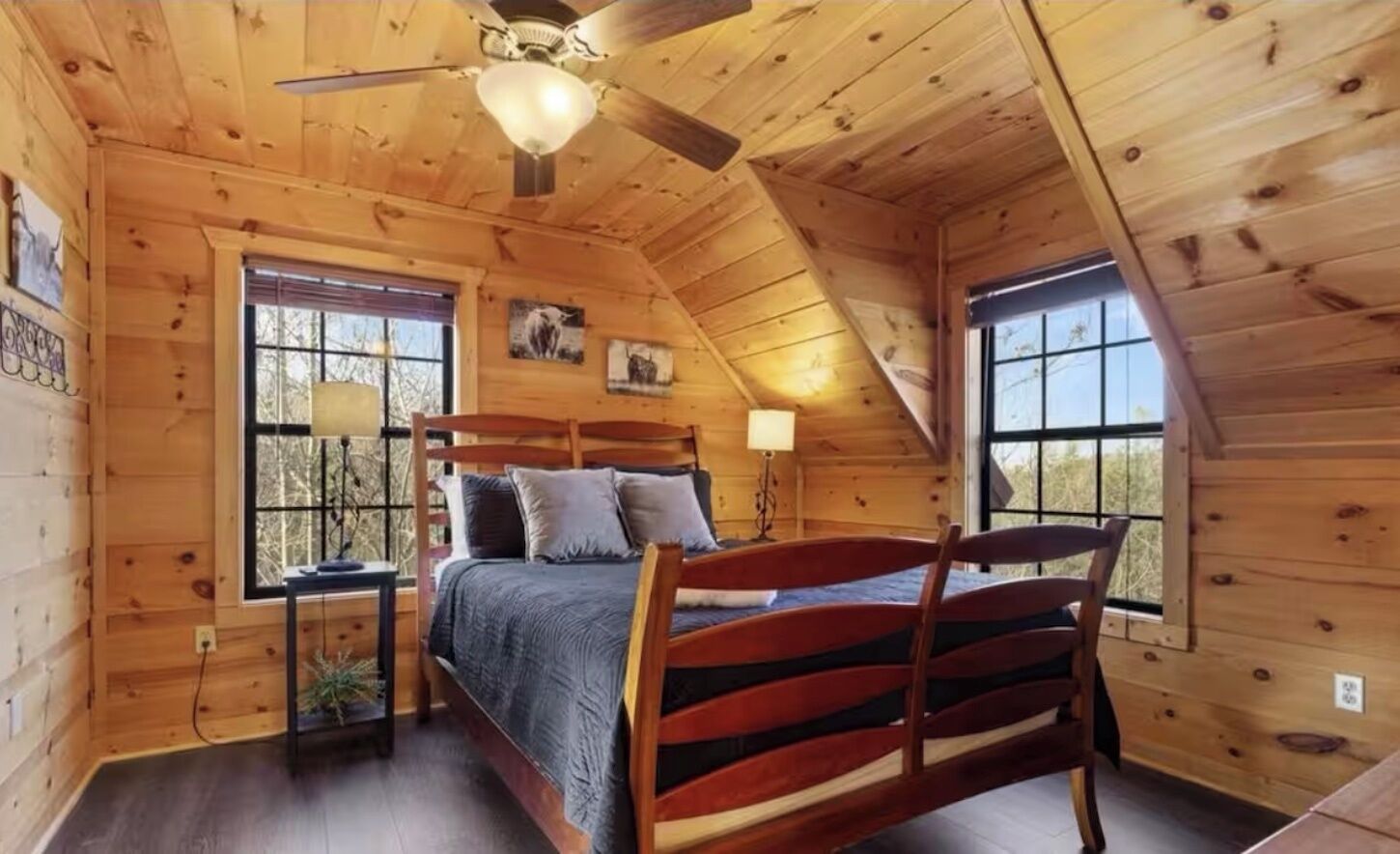 Une chambre à coucher située dans une cabane en bois, comprenant un lit, deux fenêtres, un ventilateur de plafond et une table de chevet.