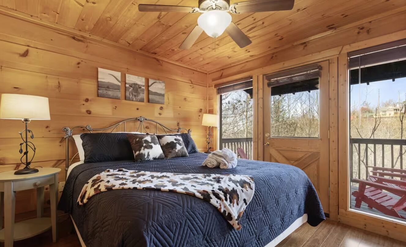 A wooden cabin bedroom with a bed, two bedside lamps, a ceiling fan, and a large window.