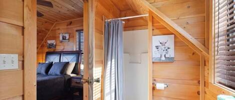 A wooden cabin with a bedroom featuring a bed, a bathroom with a shower curtain, and a small window with blinds.
