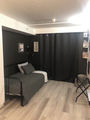 Quarto pequeno com cama de solteiro, roupa de cama escura, cortina preta, luminária no teto e prateleira com objetos decorativos.