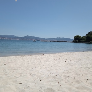 A beach with clear blue water, a sandy shore, and a distant cityscape with mountains.