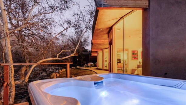 A hot tub area with a wooden deck, a stone wall, and a glass door leading to a room with a bed and a chair. - River Spirit- Arquitectural wonder of the Sequoias (Three Rivers)