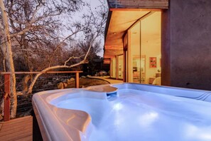 A hot tub area with a wooden deck, a stone wall, and a glass door leading to a room with a bed and a chair. - River Spirit- Arquitectural wonder of the Sequoias (Three Rivers)