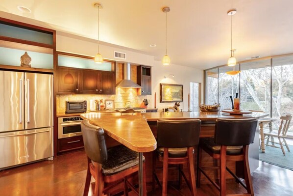 A modern kitchen with a central island, bar stools, stainless steel appliances, and large windows offering a view. - River Spirit- Arquitectural wonder of the Sequoias (Three Rivers)