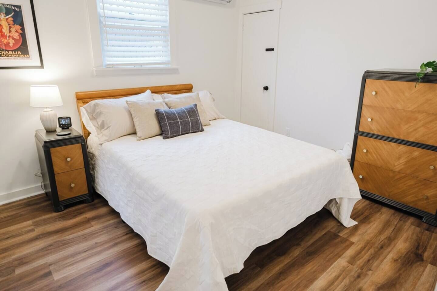 Un dormitorio ordenado con cama, mesita de noche, cómoda y ventana con persianas.