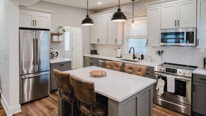 A modern kitchen with stainless steel appliances, a central island with bar stools, pendant lighting, and a view of the outdoors through a window.