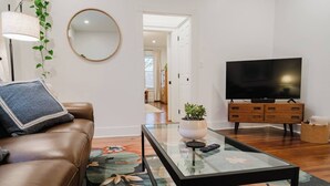 A living room with a brown leather sofa, a glass coffee table with a plant, a flat-screen TV on a wooden stand, and a large mirror on the wall.