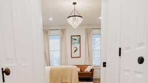 A hallway leading to a bedroom with a wooden floor, a bed with a white blanket, a chandelier, and a framed picture on the wall.