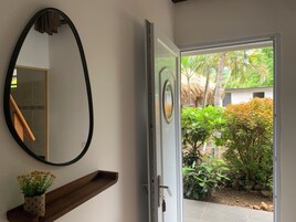 Una habitación con un espejo, un estante de madera con una planta en maceta y una puerta que da a un jardín con plantas tropicales.